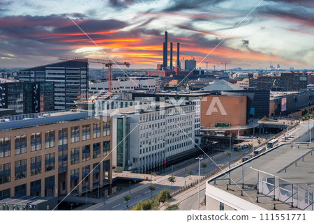 Capturing a Scandinavian cityscape at sunrise or sunset, this image features modern buildings with a mix of designs, construction cranes, and industrial chimneys in Copenhagen. 111551771