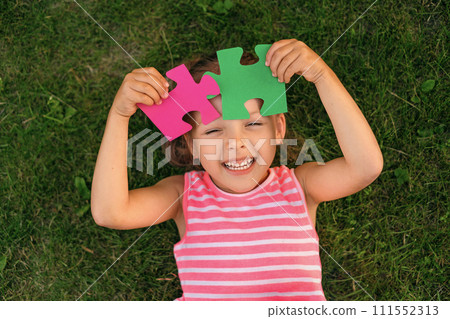 Funny laughing autistic girl playing with colored puzzle pieces lying on the grass in the garden Funny laughing autistic girl playing with colored puzzle pieces lying on the grass in the garden 111552313