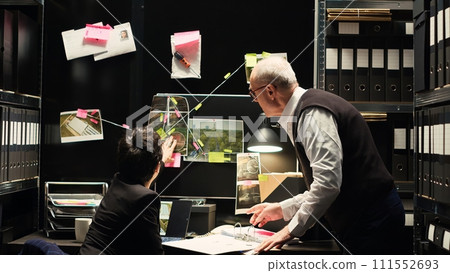 Partners gather criminal intelligence on clues board, teamwork to solve mistery and catch culprit. Experienced criminologist and police officer discussing about investigation report. Tripod shot. Partners gather criminal intelligence on clues board, teamwork to solve mistery and catch culprit. Experienced criminologist and police officer discussing about investigation report. Tripod shot. 111552693