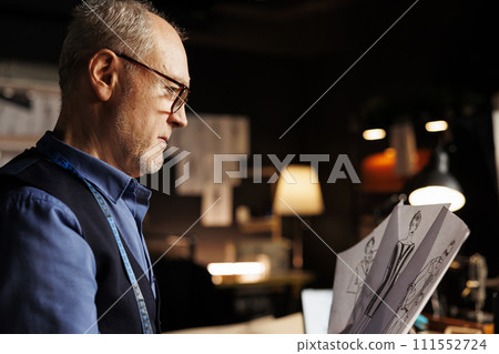 Close up of elderly suitmaker comparing handdrawn sketch designs, trying to pick perfect attire for upcoming runway show. Senior couturier in tailoring studio manufacturing premium collection Close up of elderly suitmaker comparing handdrawn sketch designs, trying to pick perfect attire for upcoming runway show. Senior couturier in tailoring studio manufacturing premium collection 111552724