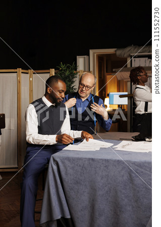 Stylish customer discussing best suit customizations options with expert suitmaker over clothing sketches in tailoring shop. Gentleman looking at fashion designs, choosing desired adjustments Stylish customer discussing best suit customizations options with expert suitmaker over clothing sketches in tailoring shop. Gentleman looking at fashion designs, choosing desired adjustments 111552730