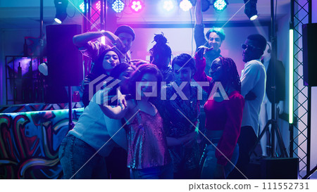 Men and women taking photos at party and dancing together under stage lights. Young adults having fun with pictures on smartphone, enjoying disco event at social gathering. Handheld shot. 111552731
