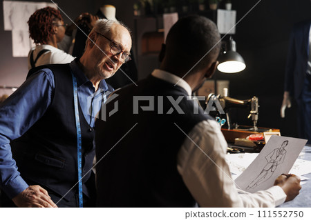 Couturiers manufacturing bespoken clothing, working together to finalize client sartorial comission. Master tailor and diverse apprentice team of suitmakers checking fashion design sketches in atelier 111552750