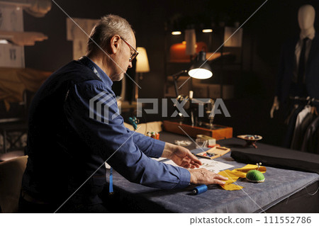 Professional elderly tailor working with sewing materials to customize bespoken sartorial garments for comissioning client in atelier shop. Senior couturier manufacturing clothing in workspace Professional elderly tailor working with sewing materials to customize bespoken sartorial garments for comissioning client in atelier shop. Senior couturier manufacturing clothing in workspace 111552786