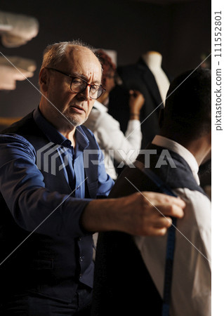 Professional expert suitmaker checking custom made suit fitting assisted by couturier. Apprentice tailor helping master fashion designer with sartorial measurements for upcoming collection, close up 111552801