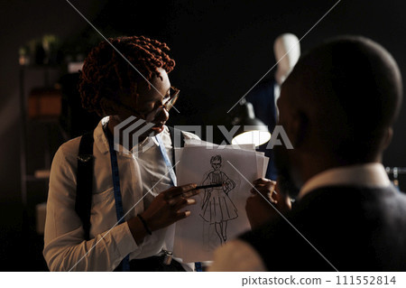 Experienced fashion designer showing couturier sketch drawing of wedding dress she wants him to customize. African american dressmakers working on stylish bespoke sartorial attire Experienced fashion designer showing couturier sketch drawing of wedding dress she wants him to customize. African american dressmakers working on stylish bespoke sartorial attire 111552814