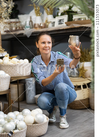 Joyful casual female comparing Christmas candle holders for home decoration in store 111553073