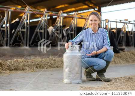 Smiling woman dairy farm worker posing with milk can in cowshed 111553074