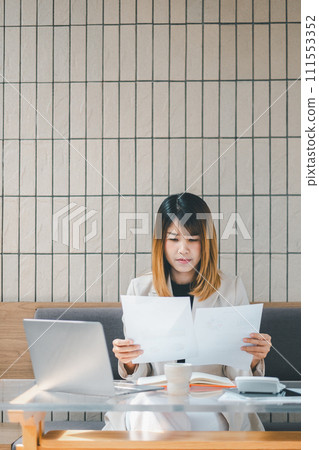 Freelancer concept, Focused businesswoman in a beige blazer reviews paperwork with her laptop at a cafe table, embodying professional multitasking. 111553352