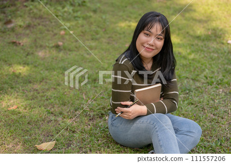 A positive young Asian woman sitting on the grass in a park with her diary. A positive young Asian woman sitting on the grass in a park with her diary. 111557206