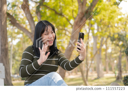 A cheerful Asian woman is talking on a video call with her friend while relaxing in a green park. 111557223