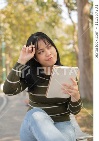 A woman is sitting on a bench in a green park with her notebook, contemplating ideas for inspiration 111557231