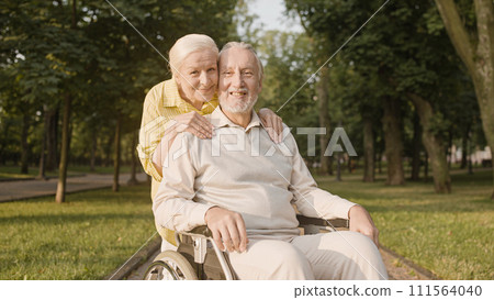 Cheerful aged wife hugging husband in wheelchair, social care, retirement home Cheerful aged wife hugging husband in wheelchair, social care, retirement home 111564040