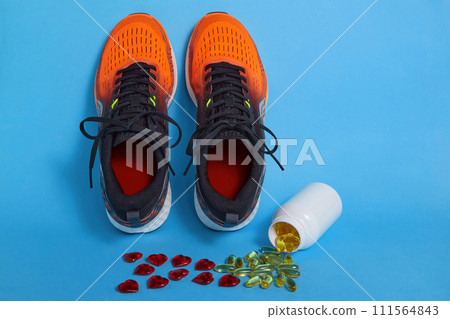 Scattered yellow vitamin D capsules from a jar and red decorative hearts and orange running sneakers on a blue background Scattered yellow vitamin D capsules from a jar and red decorative hearts and orange running sneakers on a blue background 111564843