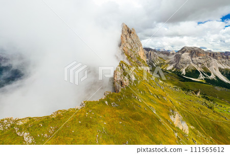 Observation point facing Seceda mountain surrounded by white dense fog. Green grass grows on slopes of rocky ridgeline aerial view 111565612