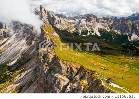 Tourist observation point against Seceda mountain with rocky peaks and green grass under cloudy sky. Scenic Italian Alps in summer aerial view Tourist observation point against Seceda mountain with rocky peaks and green grass under cloudy sky. Scenic Italian Alps in summer aerial view 111565633