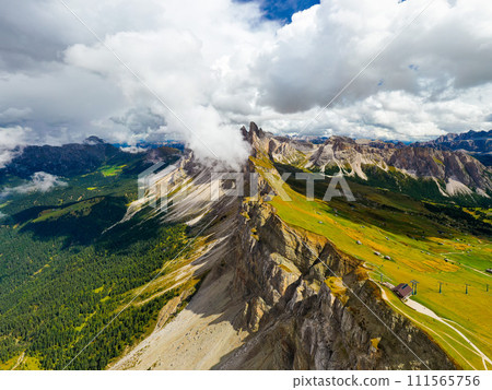 Fresh green grass grows in valley at Seceda ridgeline foot in Italian Alps. Wild nature and rocky mountain in highland on cloudy day aerial view 111565756