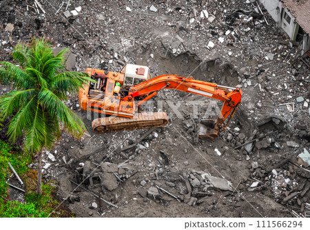 An excavator clears ruins next to a surviving palm tree. Aerial view of demolition site with ruined old building. Destruction of dilapidated housing for new development. An excavator clears ruins next to a surviving palm tree. Aerial view of demolition site with ruined old building. Destruction of dilapidated housing for new development. 111566294