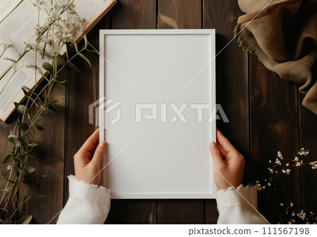 Girl holds a white poster with white frame mockup in her hands with green plants around on wooden background Girl holds a white poster with white frame mockup in her hands with green plants around on wooden background 111567198