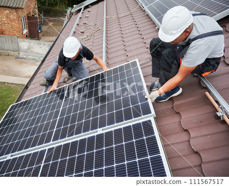 Men technicians mounting photovoltaic solar moduls on roof of house. Builders in helmets installing solar panel system outdoors. Concept of alternative and renewable energy. 111567517