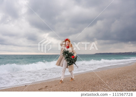 Redhead woman Christmas tree sea. Christmas portrait of a happy redhead woman walking along the beach and holding a Christmas tree in her hands. Dressed in a light coat, white suit and red mittens. Redhead woman Christmas tree sea. Christmas portrait of a happy redhead woman walking along the beach and holding a Christmas tree in her hands. Dressed in a light coat, white suit and red mittens. 111567728