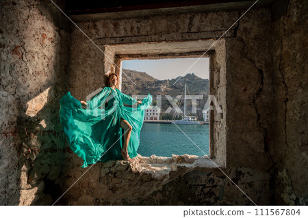 Rear view of a happy blonde woman in a long mint dress posing against the backdrop of the sea in an old building with columns. Girl in nature against the blue sky. 111567804