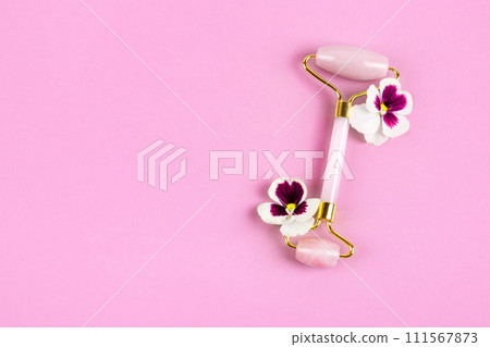 Pink jade roller for face massage with flowers pansies. Facial massager tools. Anti age, lifting and toning care. Modern selfcare concept. Pink jade roller for face massage with flowers pansies. Facial massager tools. Anti age, lifting and toning care. Modern selfcare concept. 111567873