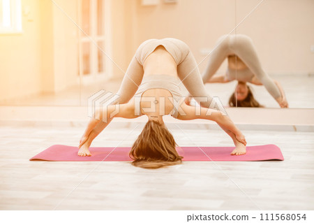 Young woman doing yoga in the gym. A girl with long hair and in a beige tracksuit stands in the Prasarita Padottanasana pose on a pink carpet. A woman performs Prasarita Padottanasana. 111568054