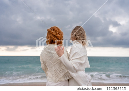 Women sea walk friendship spring. Two girlfriends, redhead and blonde, middle-aged walk along the sandy beach of the sea, dressed in white clothes. Against the backdrop of a cloudy sky and the winter Women sea walk friendship spring. Two girlfriends, redhead and blonde, middle-aged walk along the sandy beach of the sea, dressed in white clothes. Against the backdrop of a cloudy sky and the winter 111568315
