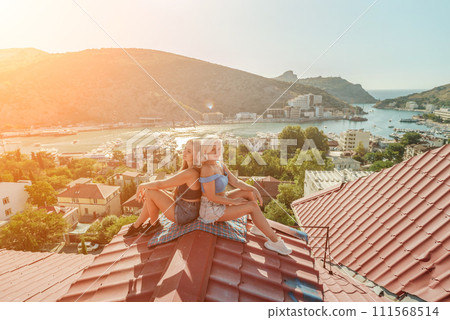 Two women sitting on a red roof, enjoying the view of the town and the sea. Rooftop vantage point. In the background, there are several boats visible on the water, adding to the picturesque scene. Two women sitting on a red roof, enjoying the view of the town and the sea. Rooftop vantage point. In the background, there are several boats visible on the water, adding to the picturesque scene. 111568514