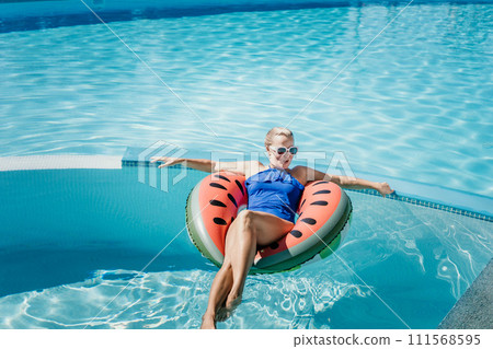Happy woman in a swimsuit and sunglasses floating on an inflatable ring in the form of a watermelon, in the pool during summer holidays and vacations. Summer concept. Happy woman in a swimsuit and sunglasses floating on an inflatable ring in the form of a watermelon, in the pool during summer holidays and vacations. Summer concept. 111568595