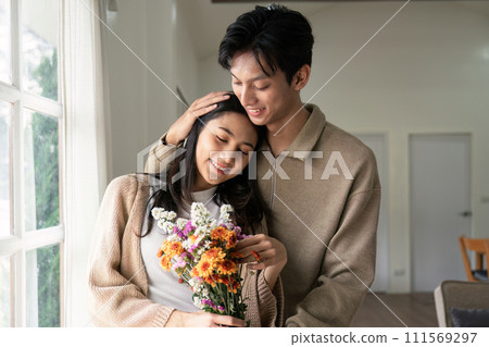 Romantic young asian couple embracing with holding flowers and smiling in living room at home. fall in love. Valentine concept 111569297