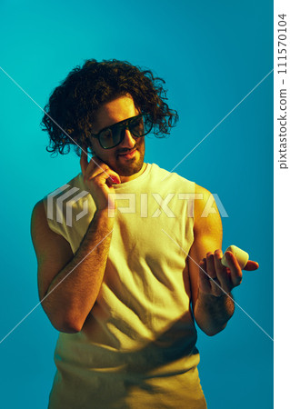 Young guy with curly messy hair and unshaved face putting earphones, listening to music against blue background in neon light Young guy with curly messy hair and unshaved face putting earphones, listening to music against blue background in neon light 111570104