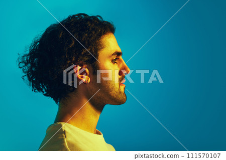 Side view portrait of young man with curly dark hair and unshaved face looking straight against blue background in neon light Side view portrait of young man with curly dark hair and unshaved face looking straight against blue background in neon light 111570107