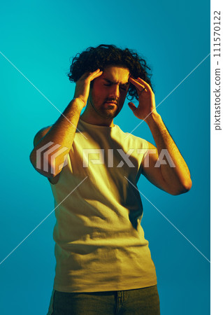 Young man in tank top holding hands on forehead, standing with eyes closed against blue background in neon light. Headache, thoughtful face 111570122