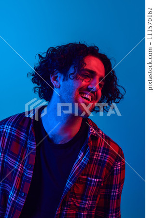 Gel portrait of young man with unshaved face, curly messy hair smiling, laughing against blue background in neon light. Joy and fun Gel portrait of young man with unshaved face, curly messy hair smiling, laughing against blue background in neon light. Joy and fun 111570162