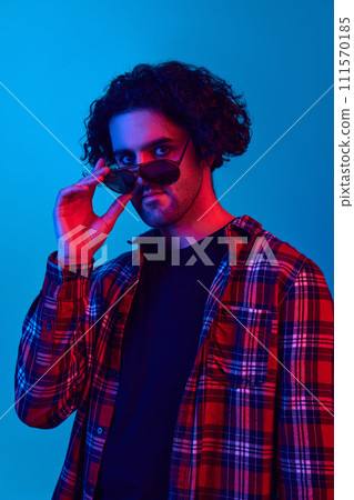 Gel portrait of young guy with curly hair, unshaved face in checkered shirt, wearing sunglasses against blue background in neon light Gel portrait of young guy with curly hair, unshaved face in checkered shirt, wearing sunglasses against blue background in neon light 111570185