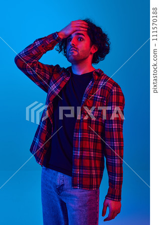 Young man wearing casual checkered shirt holding hand on forehead, expressing shocked expression against blue background in neon light Young man wearing casual checkered shirt holding hand on forehead, expressing shocked expression against blue background in neon light 111570188