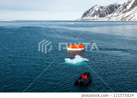 Maneuvering orange lifeboat in water in Arctic waters, Svalbard. Abandon ship drill. Lifeboat training. Man over board drill. 111573824