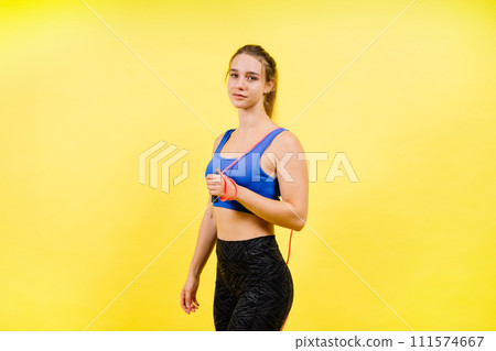 Portrait of gentle muscular woman holding skipping rope on her neck isolated over yellow background Portrait of gentle muscular woman holding skipping rope on her neck isolated over yellow background 111574667