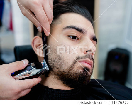 A man sitting in a barbers chair having his beard trimmed A man sitting in a barbers chair having his beard trimmed 111576272