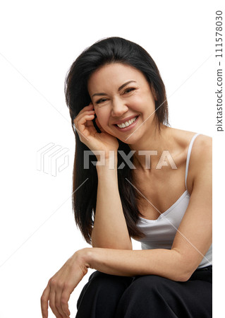 Portrait of happy, smiling, middle aged woman in white tank top smiling, seated against white studio background. 111578030