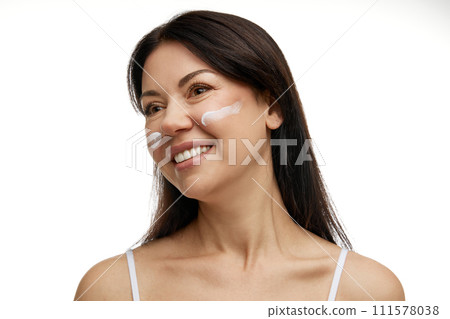 Pretty middle-aged woman with swatches of cream and looking away against white studio background. Self care treatment and protection of skin. 111578038