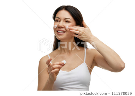 Portrait of happy, middle aged, brunette woman applying SPF cream to protect her skin in summer against white studio background. 111578039