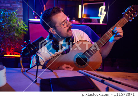 Portrait of joyful musician in glasses recording music, sings playing guitar in colorful studio, home-office. 111578071