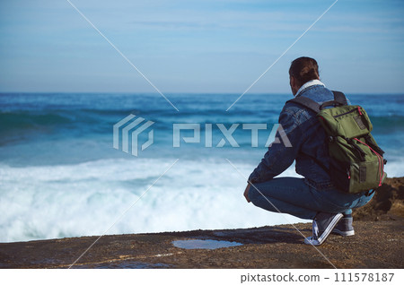Young male traveler with backpack, admiring the dangerous splashing waves from rocky cliff. People and nature. Ad space Young male traveler with backpack, admiring the dangerous splashing waves from rocky cliff. People and nature. Ad space 111578187