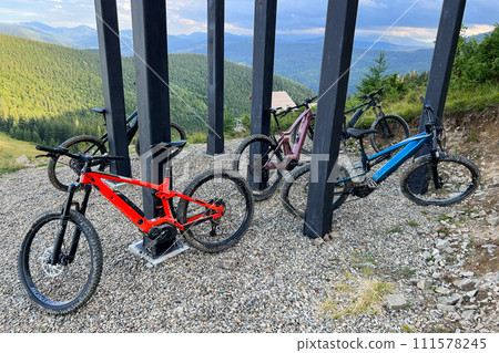 Collection of mountain electric bikes parked on a gravel-covered ground in the mountains. MTB bikes are placed under a structure with thick black pillars. 111578245