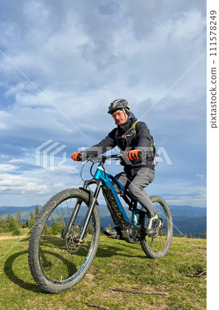 Cyclist man riding electric mountain bike outdoors. Male tourist biking along grassy trail in the mountains, wearing helmet and backpack. Concept of sport, active leisure and nature. Cyclist man riding electric mountain bike outdoors. Male tourist biking along grassy trail in the mountains, wearing helmet and backpack. Concept of sport, active leisure and nature. 111578249