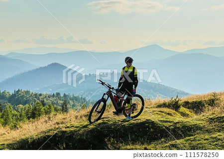 Cyclist man riding electric bike outdoors on sunny day. Male tourist resting on grassy hill, enjoying beautiful mountain landscape, wearing helmet and backpack. Concept of active leisure. Cyclist man riding electric bike outdoors on sunny day. Male tourist resting on grassy hill, enjoying beautiful mountain landscape, wearing helmet and backpack. Concept of active leisure. 111578250