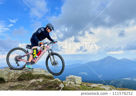 Cyclist man riding electric mountain bike outdoors. Male tourist biking along grassy trail in the mountains, wearing helmet. Concept of sport, active leisure and nature. Cyclist man riding electric mountain bike outdoors. Male tourist biking along grassy trail in the mountains, wearing helmet. Concept of sport, active leisure and nature. 111578252